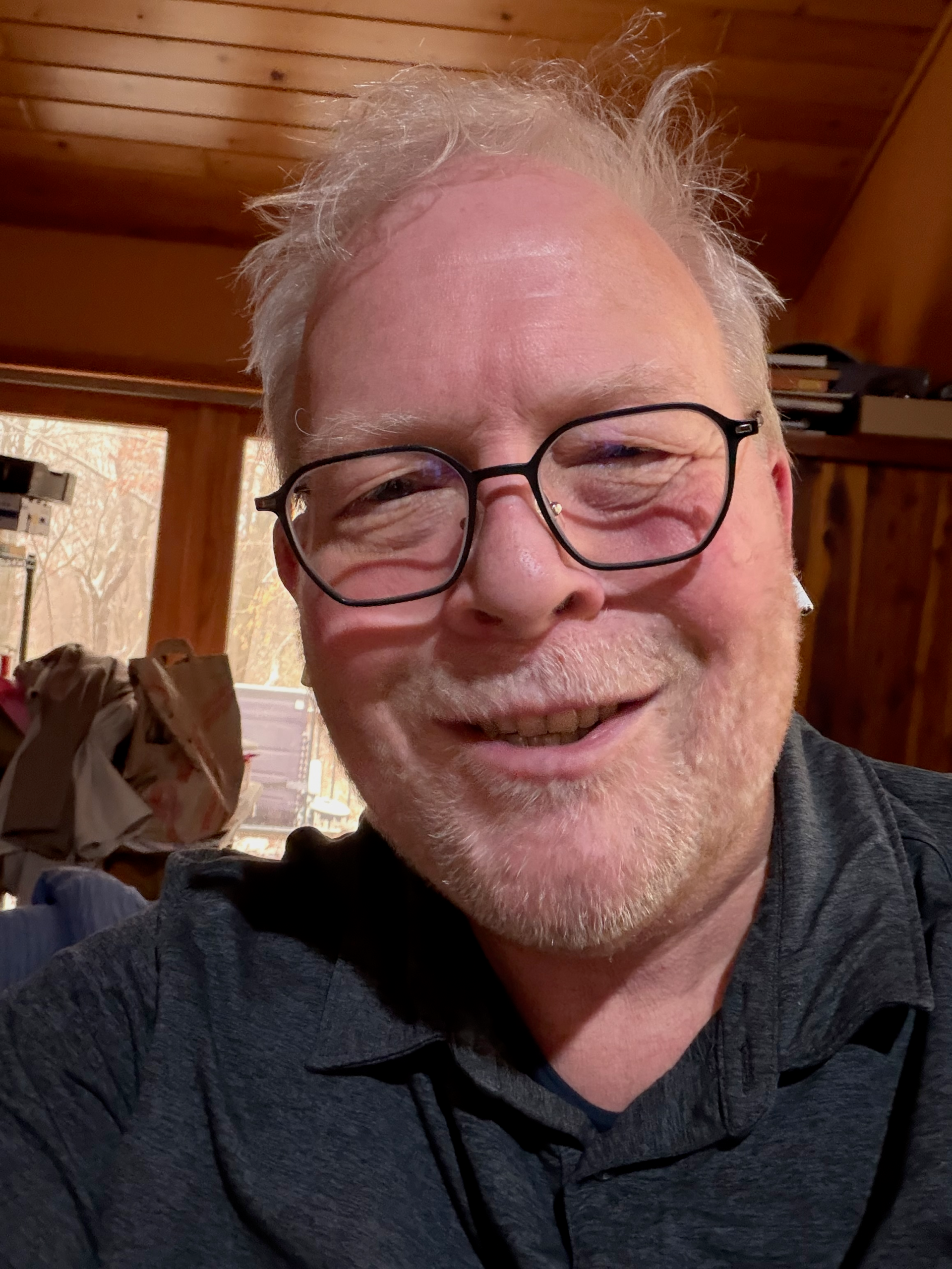 Tim, smiling wearing a dark polo shirt and dark glasses. His blondish-grey hair is short and messy. In the background, a wooden wardrobe with books stacked on top and glass doors with a view of a wooded area outside. There is a sloping ceiling that is also made from wood.